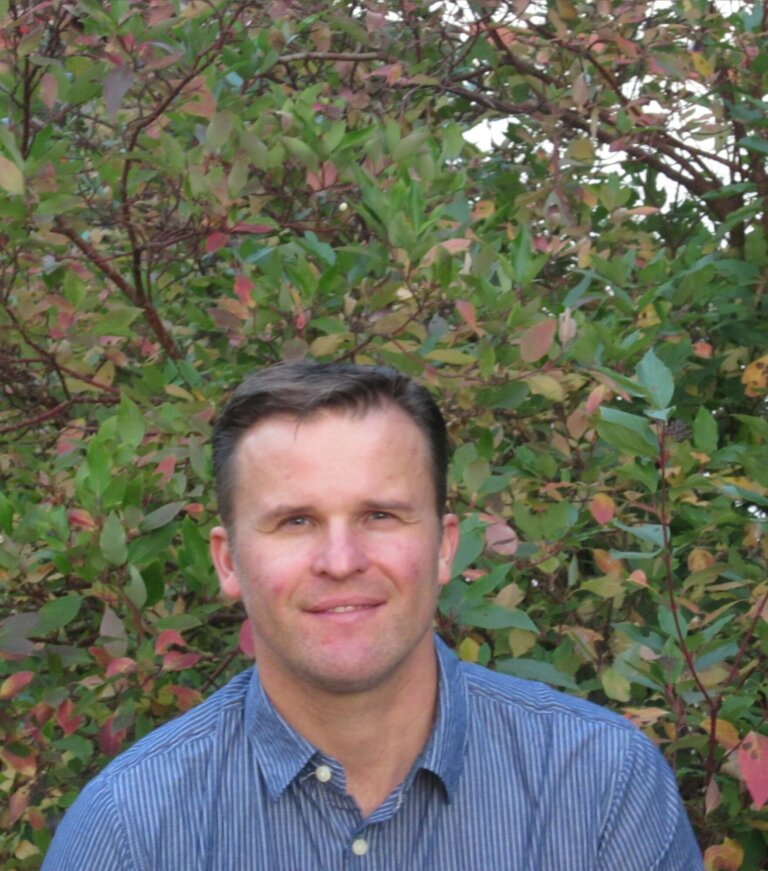 A man wearing a blue button down shirt and neat hair in front of some foliage.