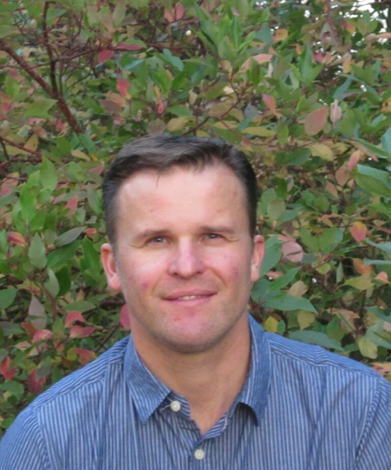 A man wearing a blue button down shirt and neat hair in front of some foliage.
